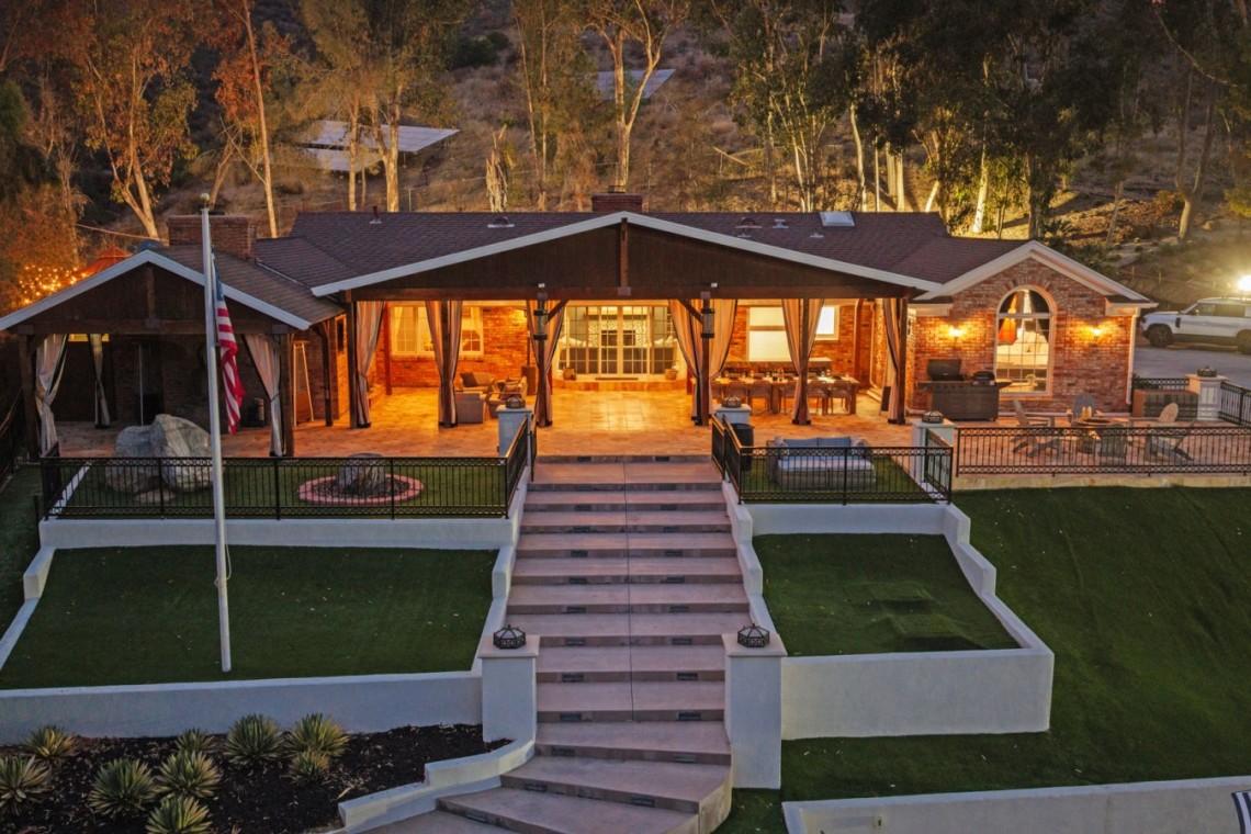 Evening view of a brick house with a large illuminated covered patio, outdoor seating, and a central stairway to a manicured lawn.