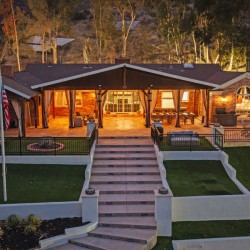 Evening view of a brick house with a large illuminated covered patio, outdoor seating, and a central stairway to a manicured lawn.