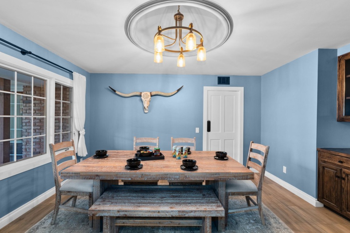 Rustic dining room with a weathered wooden table, bench, and chairs, blue walls, a longhorn skull wall art, and a brass chandelier.