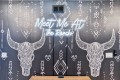 Wall mural with two white bull skulls and tribal patterns on a dark backdrop, centered neon sign reading 'Meet Me At The Ranch'.