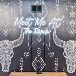 Wall mural with two white bull skulls and tribal patterns on a dark backdrop, centered neon sign reading 'Meet Me At The Ranch'.
