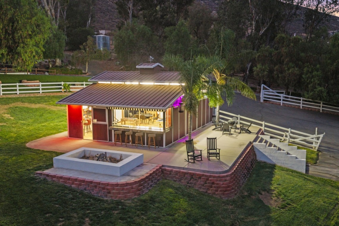 Single-story metal-roof building with an outdoor bar and stools, lit up at dusk on a landscaped yard with a palm tree nearby.