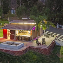 Single-story metal-roof building with an outdoor bar and stools, lit up at dusk on a landscaped yard with a palm tree nearby.