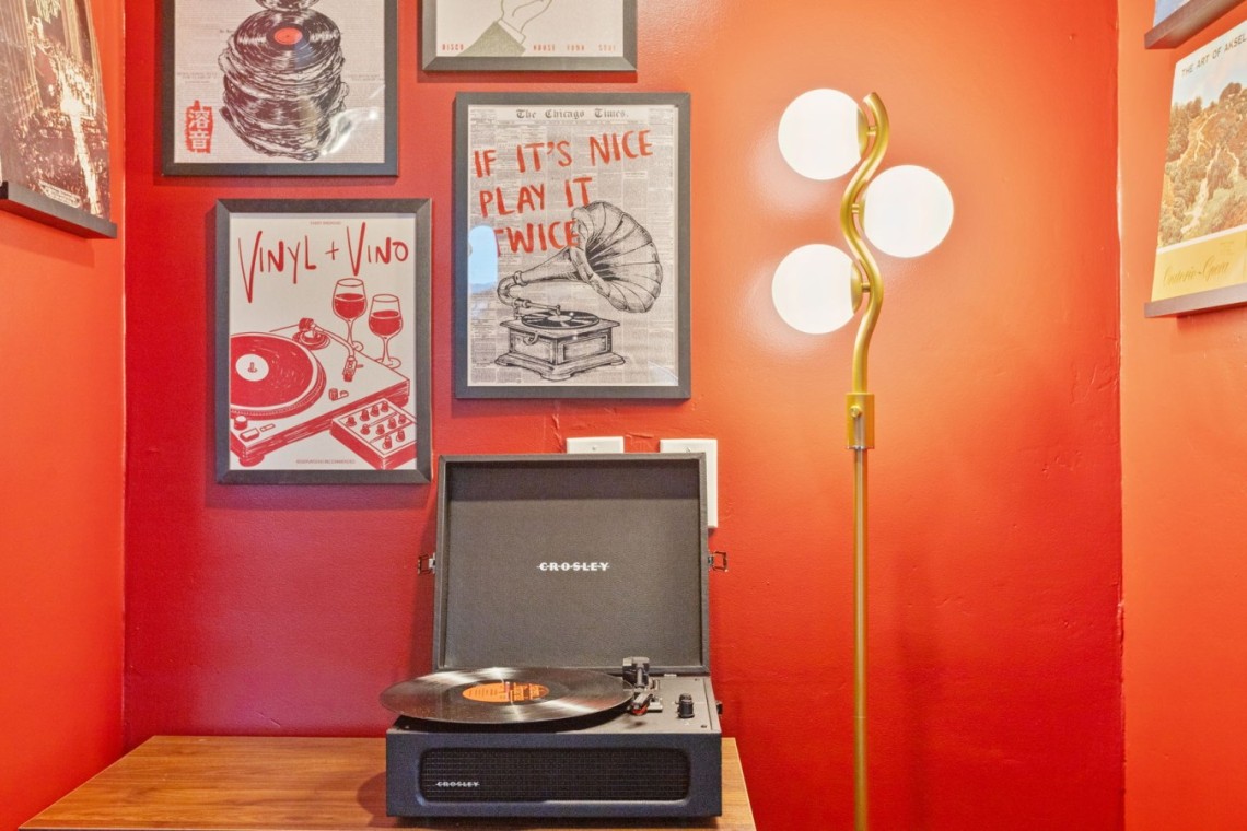 Turntable on a wooden table against a red wall with framed retro posters and a tall gold floor lamp nearby.