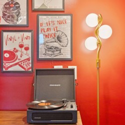 Turntable on a wooden table against a red wall with framed retro posters and a tall gold floor lamp nearby.