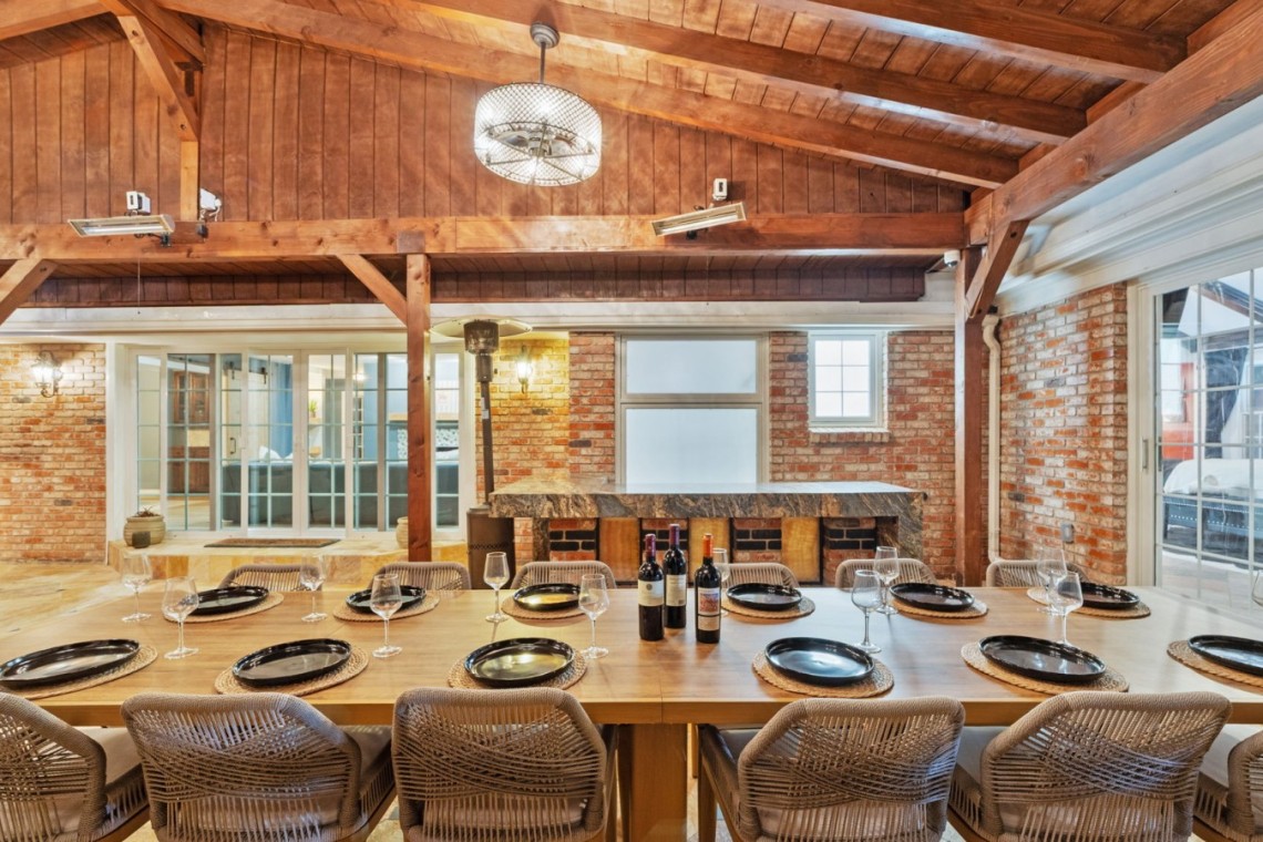 Rustic brick dining room with a long wooden table set for a meal, wine bottles in the center and woven chairs around it.
