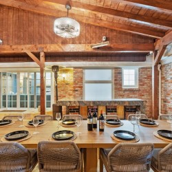 Rustic brick dining room with a long wooden table set for a meal, wine bottles in the center and woven chairs around it.