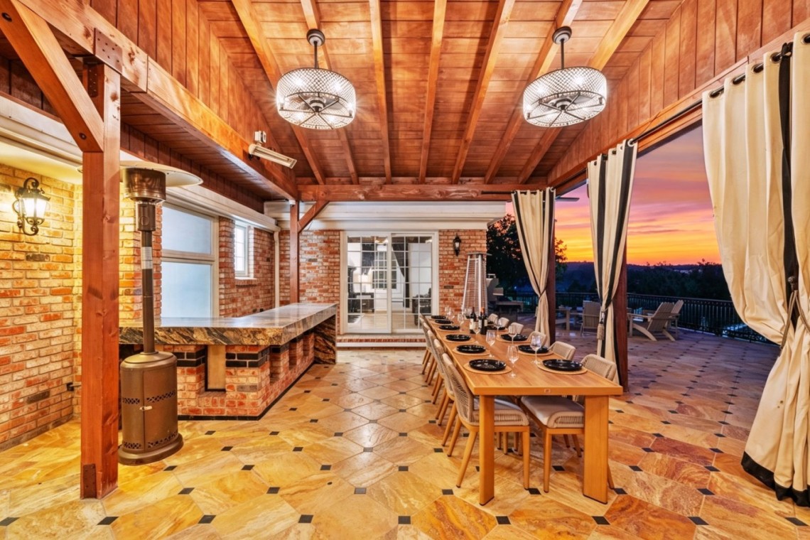 Outdoor covered patio with brick walls and a long dining table set for 12, facing a sunset view on the horizon.