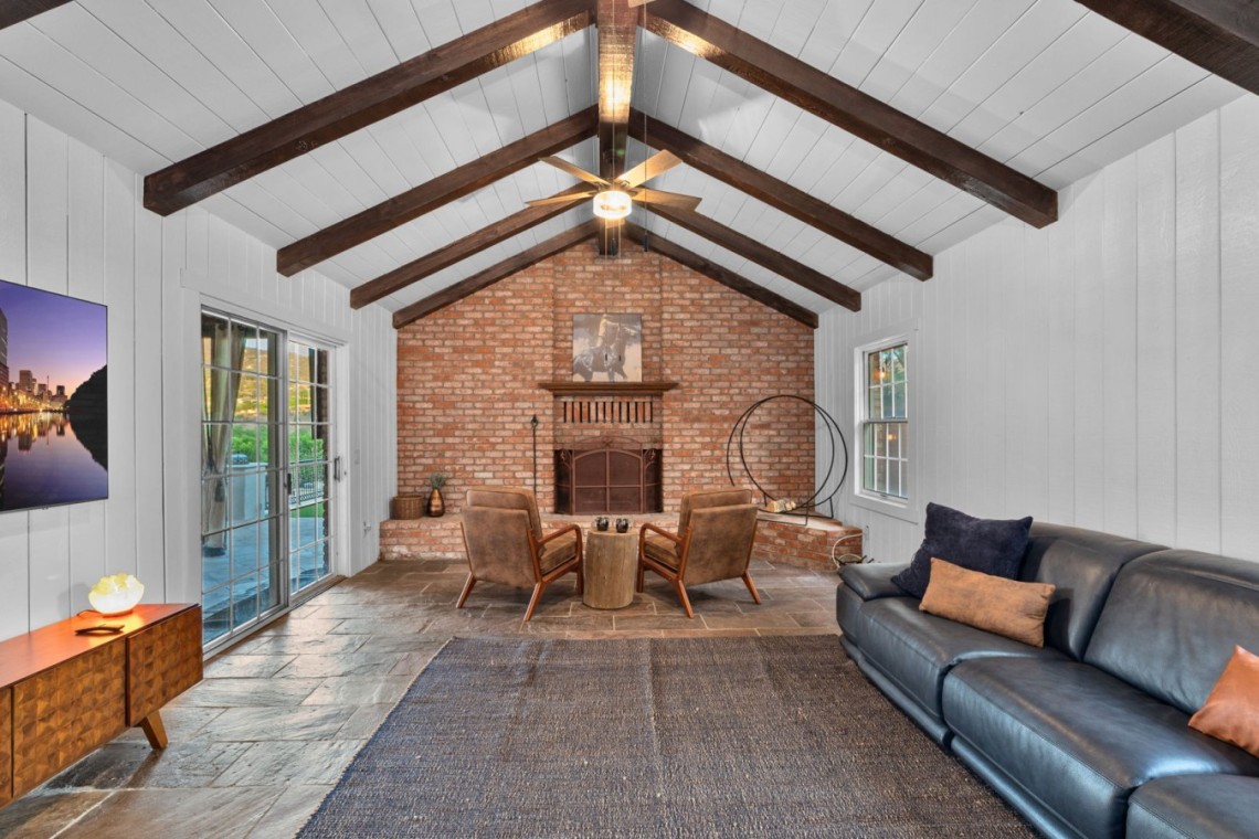 Cozy living room with a brick fireplace, vaulted ceiling with exposed dark wood beams, and a sliding glass door outside.