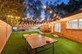 Backyard at dusk with string lights, a ping-pong table, hammock, and a brick house along a wooden fence.