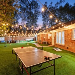Backyard at dusk with string lights, a ping-pong table, hammock, and a brick house along a wooden fence.