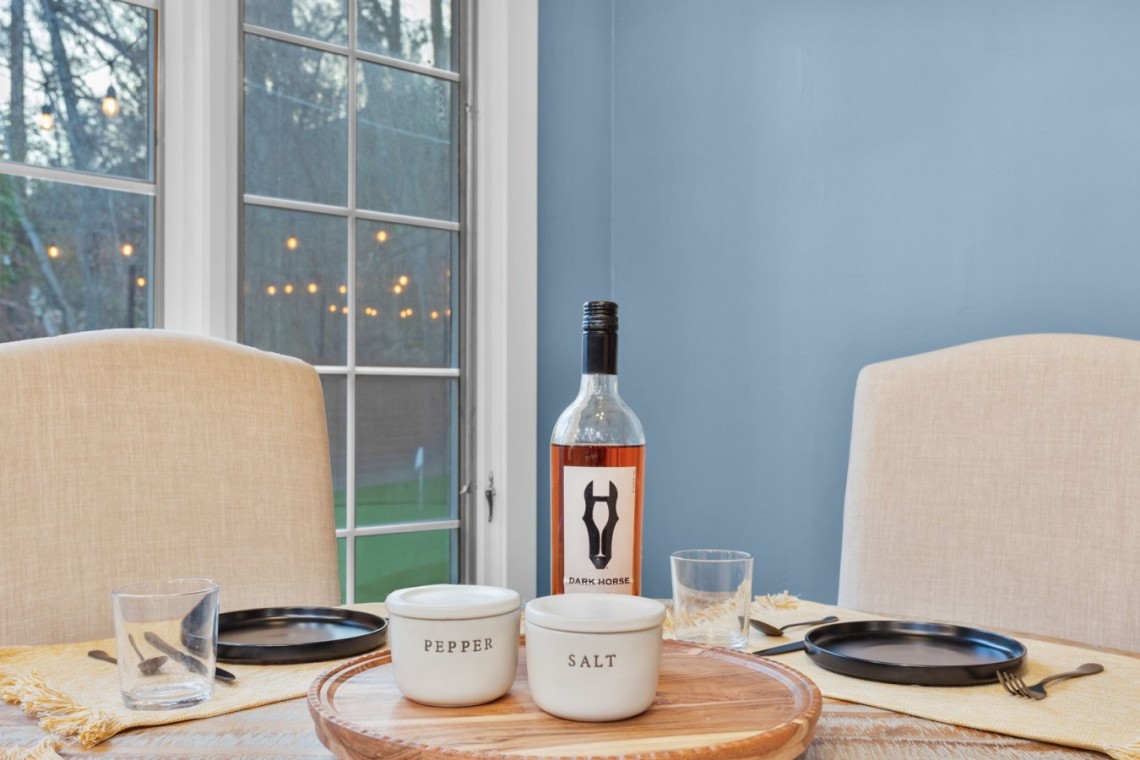 Dining table setup with a wine bottle, salt and pepper bowls, and empty glasses against a blue wall and beige chairs.