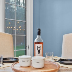 Dining table setup with a wine bottle, salt and pepper bowls, and empty glasses against a blue wall and beige chairs.