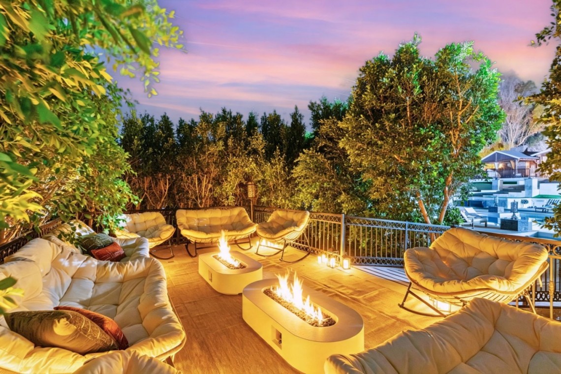 Cozy rooftop patio with cream cushioned seating around two fire pits at dusk, surrounded by greenery and lanterns