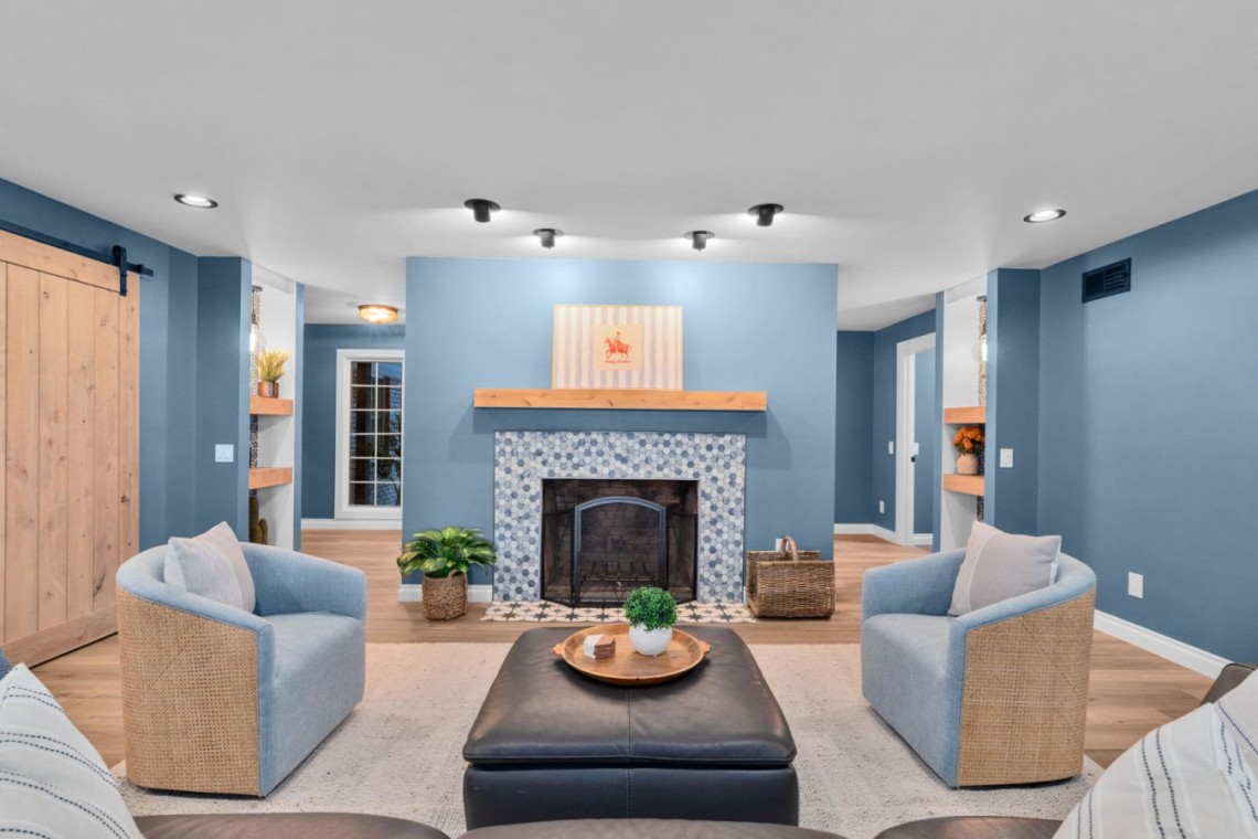Cozy living room with blue walls, a mosaic-tile fireplace, wooden mantel, and blue-and-wicker chairs arranged around a central ottoman.