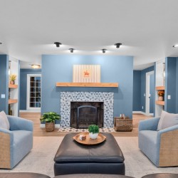 Cozy living room with blue walls, a mosaic-tile fireplace, wooden mantel, and blue-and-wicker chairs arranged around a central ottoman.