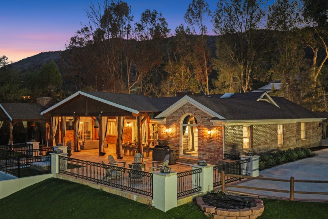 Luxurious backyard of a brick house at dusk with a lit covered outdoor living area and a stone patio, surrounded by a black metal fence and hills in the background.