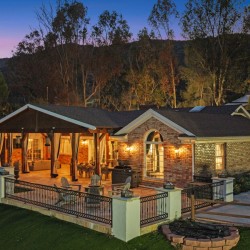 Luxurious backyard of a brick house at dusk with a lit covered outdoor living area and a stone patio, surrounded by a black metal fence and hills in the background.