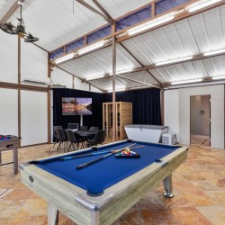Bright game room with a blue pool table in the foreground, foosball, a dining table, and a TV area in back.