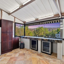 Modern kitchen with a stainless steel French-door fridge, dark wood cabinets, and marble countertop, overlooking a sunset hillside from a large window.