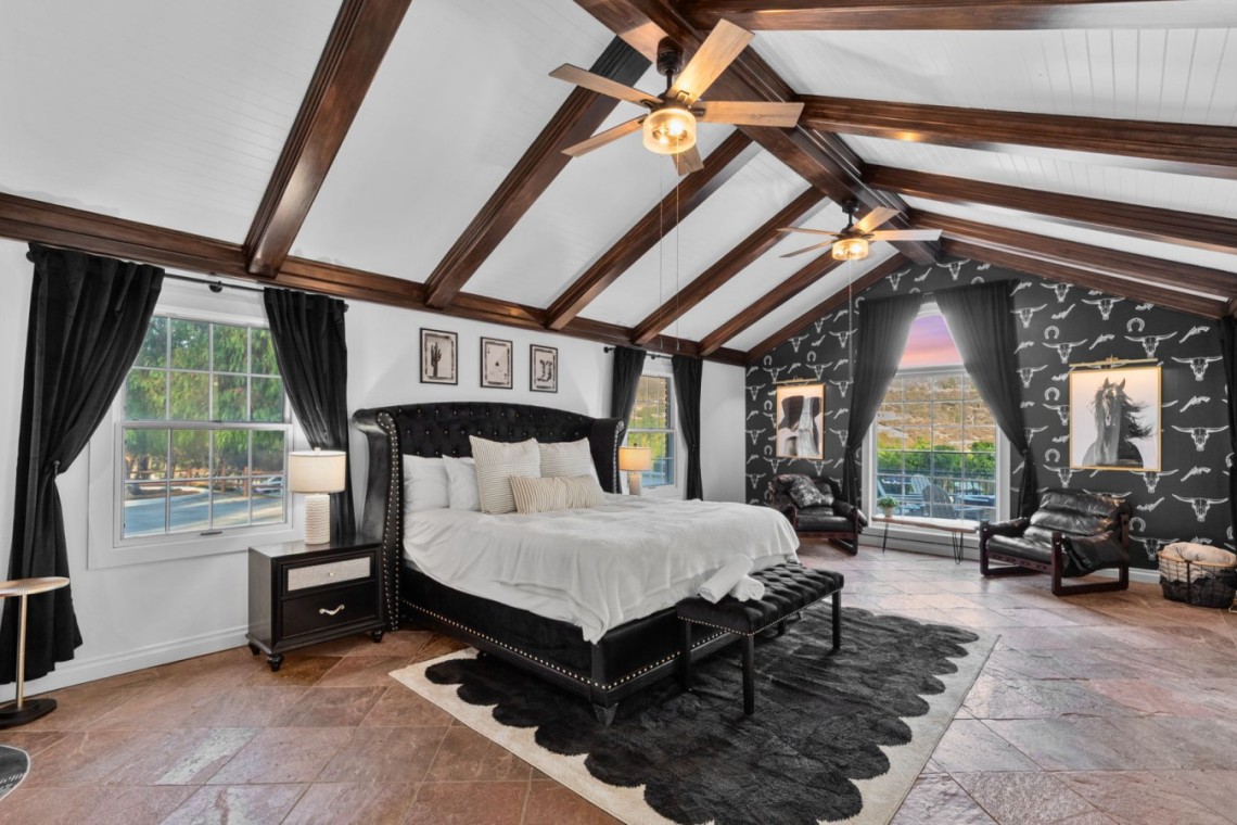Luxurious bedroom with vaulted white ceiling, dark wood beams, and a black tufted bed with matching nightstands.
