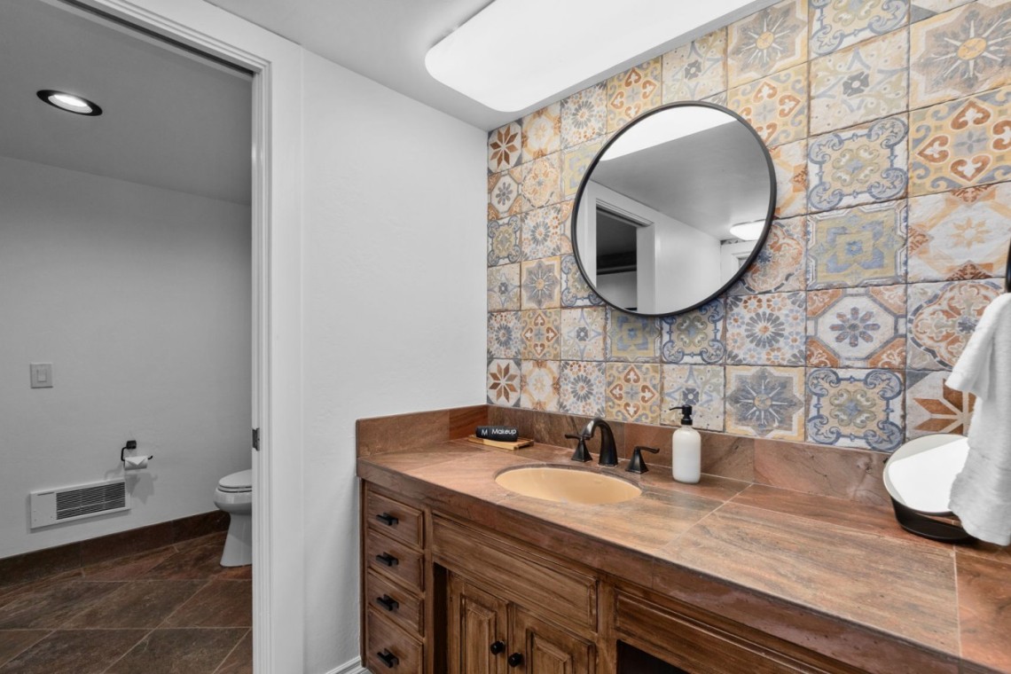 Bathroom vanity with round wall mirror, colorful patterned tile backsplash, and brown stone countertop; soap dispenser by the sink and a towel on a hook nearby.