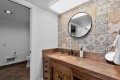 Bathroom vanity with round wall mirror, colorful patterned tile backsplash, and brown stone countertop; soap dispenser by the sink and a towel on a hook nearby.