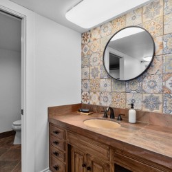Bathroom vanity with round wall mirror, colorful patterned tile backsplash, and brown stone countertop; soap dispenser by the sink and a towel on a hook nearby.