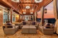 Outdoor covered lounge with wicker sectional seating, beige cushions, and purple throw pillows under a wooden beam ceiling at sunset.
