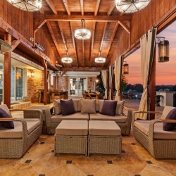 Outdoor covered lounge with wicker sectional seating, beige cushions, and purple throw pillows under a wooden beam ceiling at sunset.