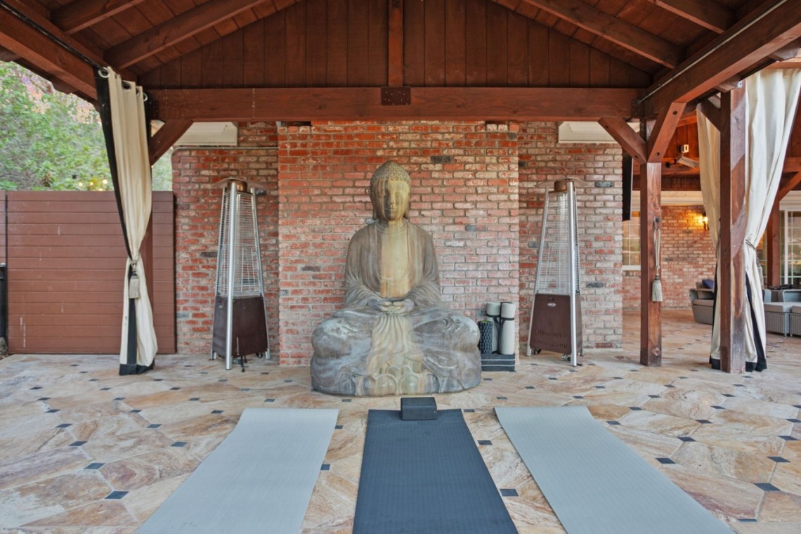 Yoga/meditation area with a large Buddha statue centered on a brick wall, three mats laid on a stone-tiled floor and cozy seating to the right under a wooden pergola.