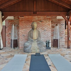Yoga/meditation area with a large Buddha statue centered on a brick wall, three mats laid on a stone-tiled floor and cozy seating to the right under a wooden pergola.
