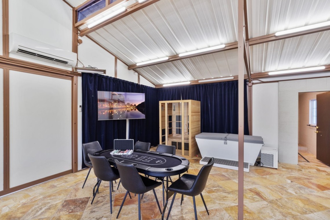 Round poker table with chips and eight chairs in a bright industrial-style room; wall TV on dark curtains, wooden sauna cabinet, and a white spa tub nearby under bright lighting.
