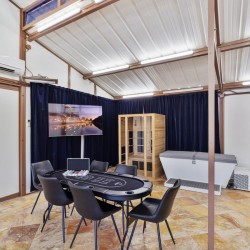 Round poker table with chips and eight chairs in a bright industrial-style room; wall TV on dark curtains, wooden sauna cabinet, and a white spa tub nearby under bright lighting.