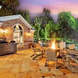 Brick house patio with outdoor grill, burning fire pit, and circular seating around the flames at sunset.