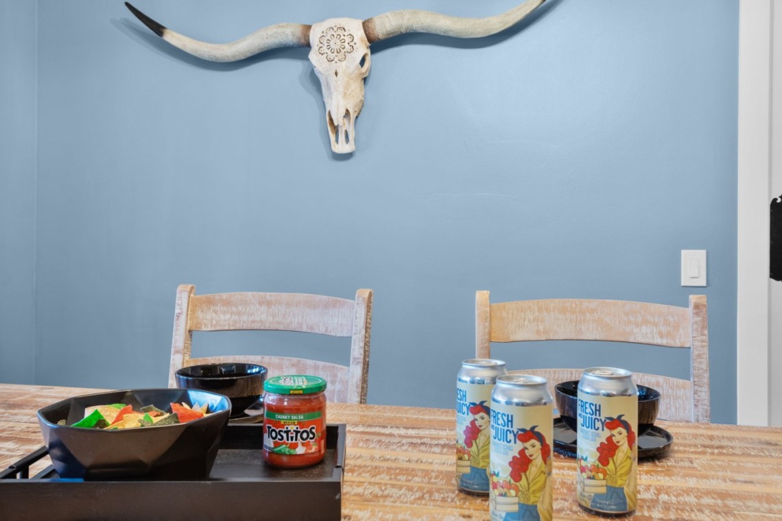Dining table with a bowl of chips, salsa jar, and four juice cans on a rustic wooden table against a blue wall with a longhorn skull mounted above.