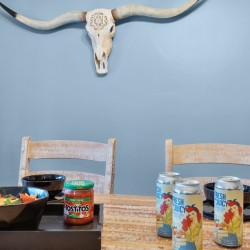 Dining table with a bowl of chips, salsa jar, and four juice cans on a rustic wooden table against a blue wall with a longhorn skull mounted above.