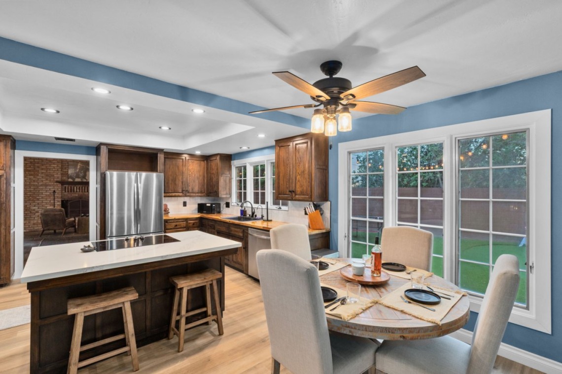 Bright blue-walled kitchen with dark wood cabinets, stainless steel refrigerator, and a central island with two stools under recessed lighting and a ceiling fan above a dining area nearby.