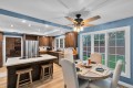 Bright blue-walled kitchen with dark wood cabinets, stainless steel refrigerator, and a central island with two stools under recessed lighting and a ceiling fan above a dining area nearby.