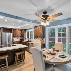 Bright blue-walled kitchen with dark wood cabinets, stainless steel refrigerator, and a central island with two stools under recessed lighting and a ceiling fan above a dining area nearby.
