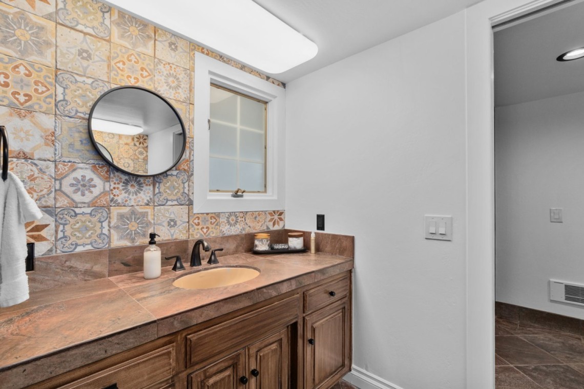 Wood-toned bathroom vanity with brown countertop, oval sink, and a colorful tile backsplash under a round mirror.