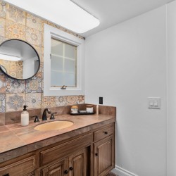 Wood-toned bathroom vanity with brown countertop, oval sink, and a colorful tile backsplash under a round mirror.