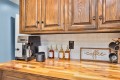 Coffee station on a wooden countertop with an espresso machine, row of syrup bottles, mug stand, and a 'coffee' sign against a blue wall.