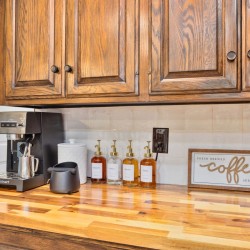 Coffee station on a wooden countertop with an espresso machine, row of syrup bottles, mug stand, and a 'coffee' sign against a blue wall.