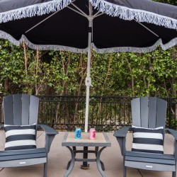 Two blue Adirondack chairs with striped pillows face a small table under a large umbrella; soda cans sit on the table in a backyard patio setting.