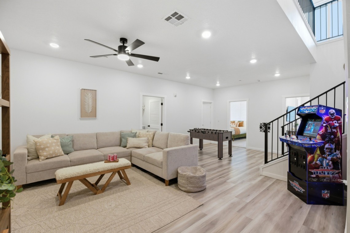 Bright open living room with a beige sectional sofa, woven bench, and neutral rug; foosball table in the back and a kids' NFL arcade machine by the stairs.