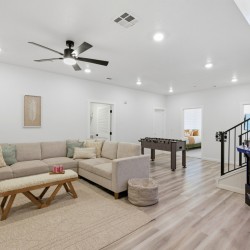 Bright open living room with a beige sectional sofa, woven bench, and neutral rug; foosball table in the back and a kids' NFL arcade machine by the stairs.