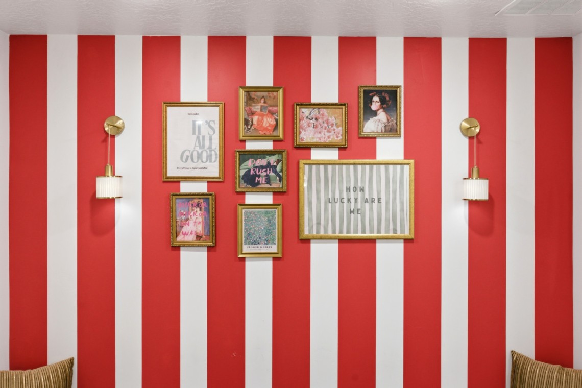 Red and white striped wall with a gallery of gold-framed artworks, including 'It’s all good' and 'How lucky are we,' flanked by brass wall sconces.