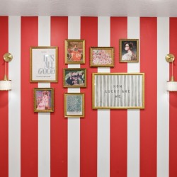 Red and white striped wall with a gallery of gold-framed artworks, including 'It’s all good' and 'How lucky are we,' flanked by brass wall sconces.