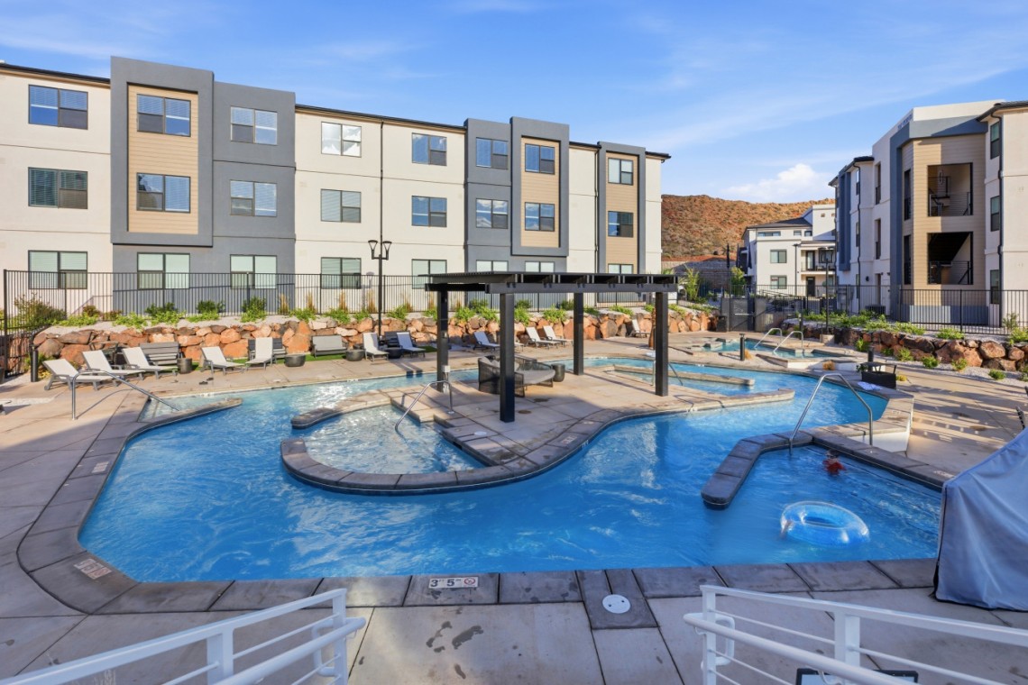 Outdoor apartment pool area with lounge chairs, a shaded seating structure, and modern beige-gray buildings nearby.